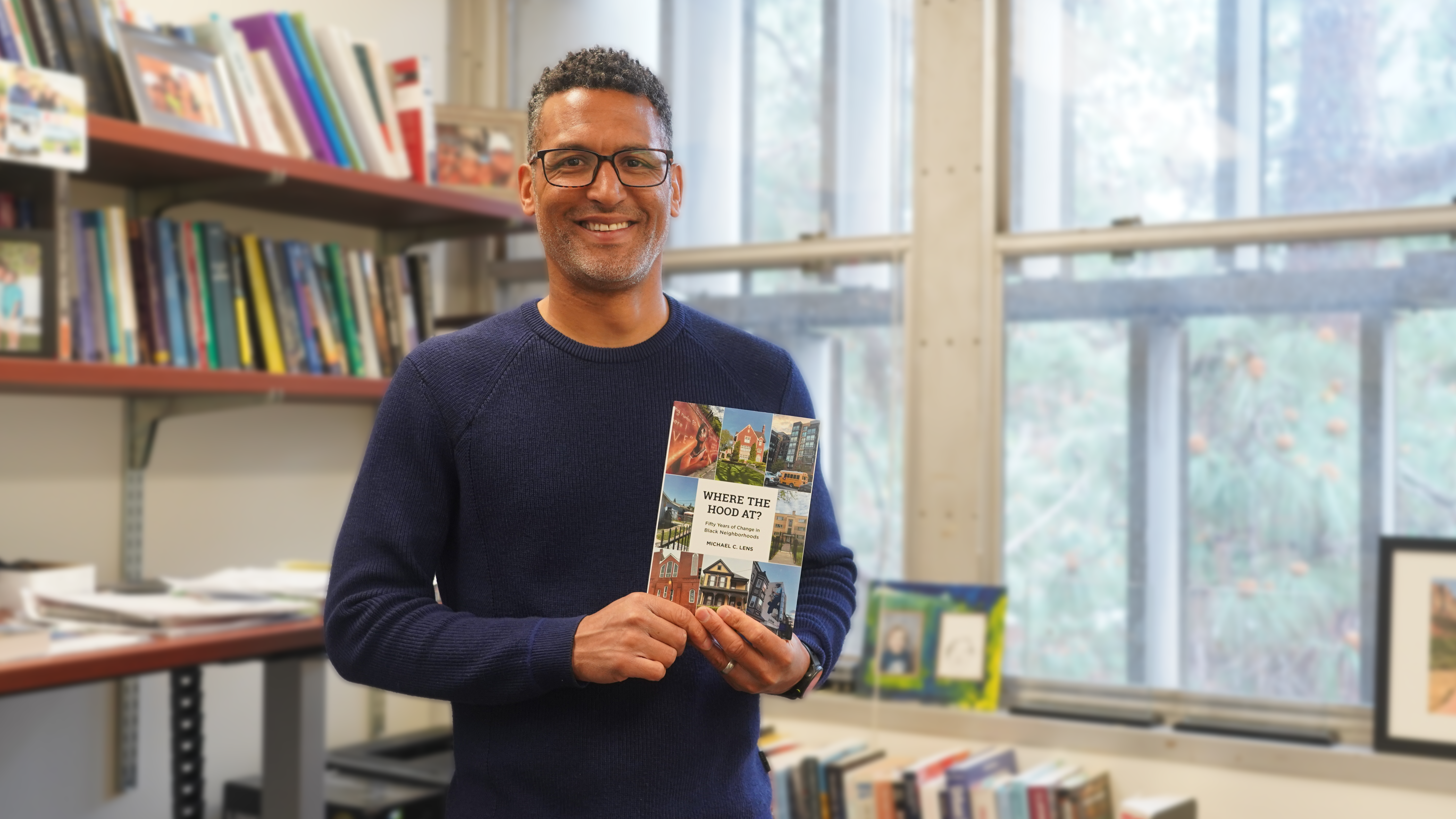 A man with glasses stands in an office filled with bookshelves, smiling while holding a book titled Where the Hood At?. The office has large windows with a view of trees outside. Shelves behind him are filled with academic books, framed photos, and office supplies